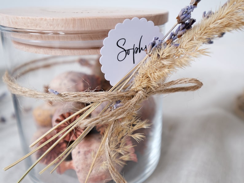 Liebevoll gestaltetes Glas mit Holzdeckel, gefüllt mit duftendem Potpourri und dekoriert mit natürlichen Gräsern und Lavendel. Perfekt als personalisiertes Geschenk oder stilvolle Deko. Der handgeschriebene Namensanhänger und die zarte Juteschnur verleihen dem Arrangement eine persönliche Note und versprühen einen Hauch von ländlicher Eleganz. Ideal für besondere Anlässe.