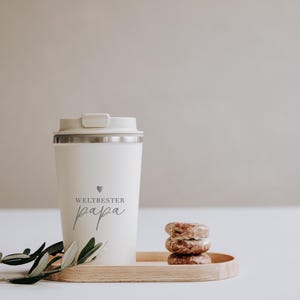 Thermobecher "Weltbester Papa" aus Edelstahl, ideal für unterwegs. Der Becher hält Getränke lange warm und ist ein perfektes Geschenk für Väter. Neben dem Becher liegt ein Holztablett mit Gebäck und einem Olivenzweig, was eine gemütliche und liebevolle Atmosphäre schafft. Perfekt für den täglichen Kaffeegenuss mit persönlichem Touch.