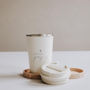 Thermobecher "Weltbester Papa" aus Edelstahl, ideal für unterwegs. Der Becher hält Getränke lange warm und ist ein perfektes Geschenk für Väter. Neben dem Becher liegt ein Holztablett mit Gebäck und einem Olivenzweig, was eine gemütliche und liebevolle Atmosphäre schafft. Perfekt für den täglichen Kaffeegenuss mit persönlichem Touch.