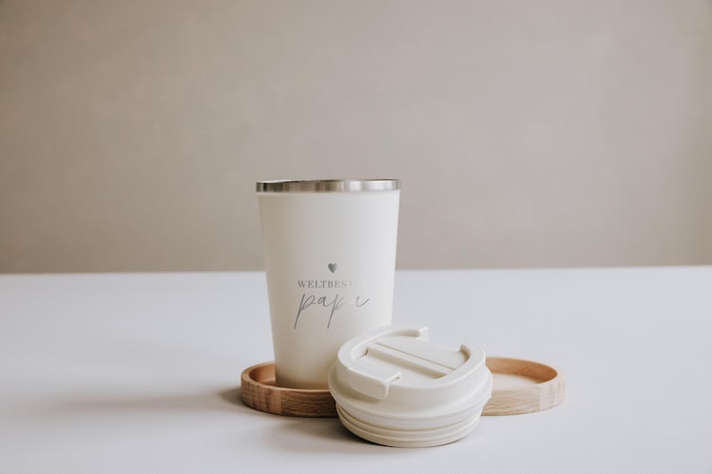 Thermobecher "Weltbester Papa" aus Edelstahl, ideal für unterwegs. Der Becher hält Getränke lange warm und ist ein perfektes Geschenk für Väter. Neben dem Becher liegt ein Holztablett mit Gebäck und einem Olivenzweig, was eine gemütliche und liebevolle Atmosphäre schafft. Perfekt für den täglichen Kaffeegenuss mit persönlichem Touch.