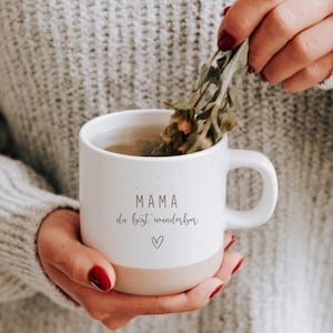 Gravierte Keramiktasse in der Hand. Tee wird in die Tasse gefüllt und sorgt für einen gemütlichen Tag. Tasse mit weißer matter Lasur und kleinen sprenkeln und unglasiertem Bodendesign in Natur, individuell anpassbar. Mama du bist wunderbar