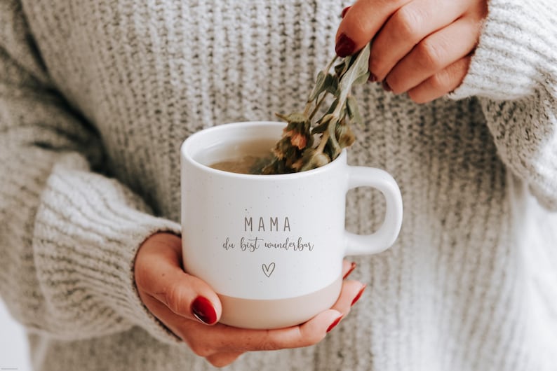 Gravierte Keramiktasse in der Hand. Tee wird in die Tasse gefüllt und sorgt für einen gemütlichen Tag. Tasse mit weißer matter Lasur und kleinen sprenkeln und unglasiertem Bodendesign in Natur, individuell anpassbar. Mama du bist wunderbar
