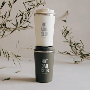 Doppelpack aus stilvollen, isolierten Bechern mit den Aufschriften "Hot Mom Club" und "Hot Dad Club" vor einem eleganten Hintergrund mit Olivenzweigen. Perfekt für Eltern, die stolz und stilvoll unterwegs sein möchten. Diese Becher halten Getränke warm und verleihen jedem Morgen einen besonderen Touch. Ideal als Geschenk oder für den eigenen täglichen Gebrauch.