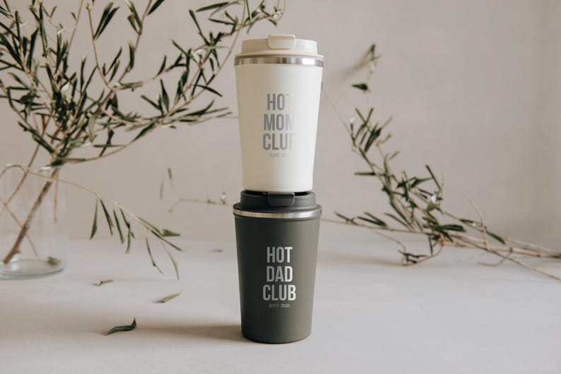 Doppelpack aus stilvollen, isolierten Bechern mit den Aufschriften "Hot Mom Club" und "Hot Dad Club" vor einem eleganten Hintergrund mit Olivenzweigen. Perfekt für Eltern, die stolz und stilvoll unterwegs sein möchten. Diese Becher halten Getränke warm und verleihen jedem Morgen einen besonderen Touch. Ideal als Geschenk oder für den eigenen täglichen Gebrauch.