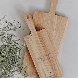 K&ouml;nnte beinhalten: Drei Holzschneidebretter mit Griffen. Die Bretter sind aus hellem Holz mit einem nat&uuml;rlichen Maserungsmuster gefertigt. Ein Brett tr&auml;gt den Text "HAND IN HAND" sowie Namen und ein Datum. Die Bretter sind in einer geschichteten Anordnung angeordnet.