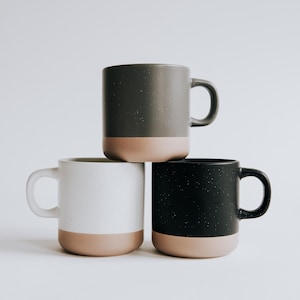 May include: Three ceramic mugs stacked on top of each other. The mugs are in shades of gray, white, and black, each with a tan-colored base. The mugs have a speckled texture and a simple, modern design.