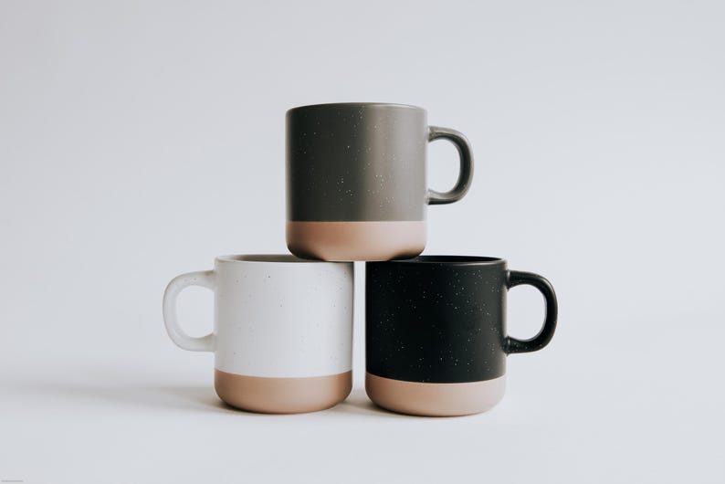 May include: Three ceramic mugs stacked on top of each other. The mugs are in shades of gray, white, and black, each with a tan-colored base. The mugs have a speckled texture and a simple, modern design.
