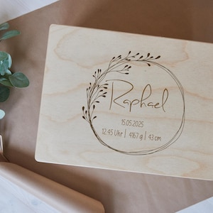 Puede incluir: Una caja de madera de color claro con texto grabado y un diseño de corona floral. El texto incluye el nombre "Raphael", la fecha, la hora, el peso (4167 g) y la altura (43 cm). La caja está sobre una superficie marrón con vegetación.