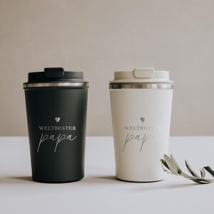 Zwei stilvolle Thermobecher in Schwarz und Weiß mit der Aufschrift „Weltbester Papa“ stehen nebeneinander auf einem hellen Tisch. Perfekt für den besten Vater der Welt, um seinen Kaffee warm zu halten. Eine zarte Olivenzweig-Dekoration rundet das elegante Design ab und verleiht dem Bild eine warme, liebevolle Note. Ideal als persönliches und praktisches Geschenk für besondere Anlässe.
