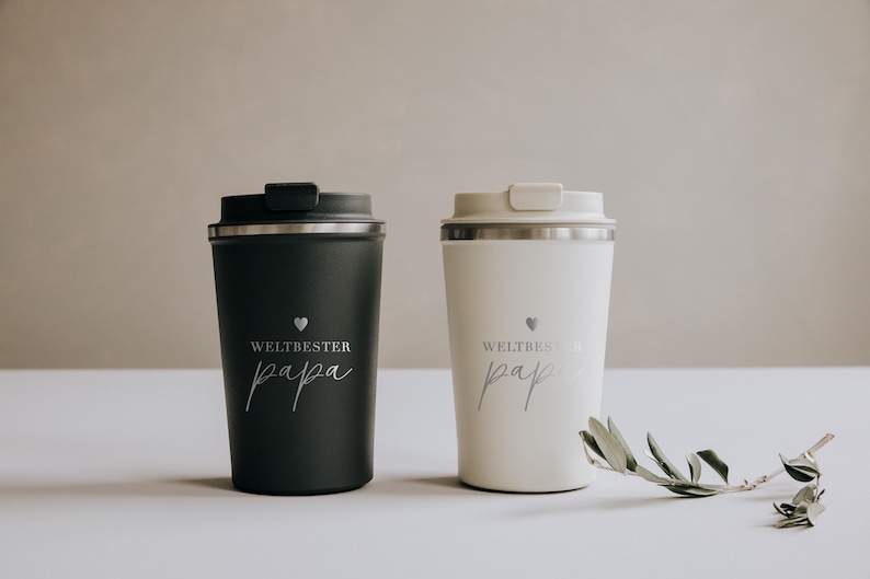Zwei stilvolle Thermobecher in Schwarz und Weiß mit der Aufschrift „Weltbester Papa“ stehen nebeneinander auf einem hellen Tisch. Perfekt für den besten Vater der Welt, um seinen Kaffee warm zu halten. Eine zarte Olivenzweig-Dekoration rundet das elegante Design ab und verleiht dem Bild eine warme, liebevolle Note. Ideal als persönliches und praktisches Geschenk für besondere Anlässe.