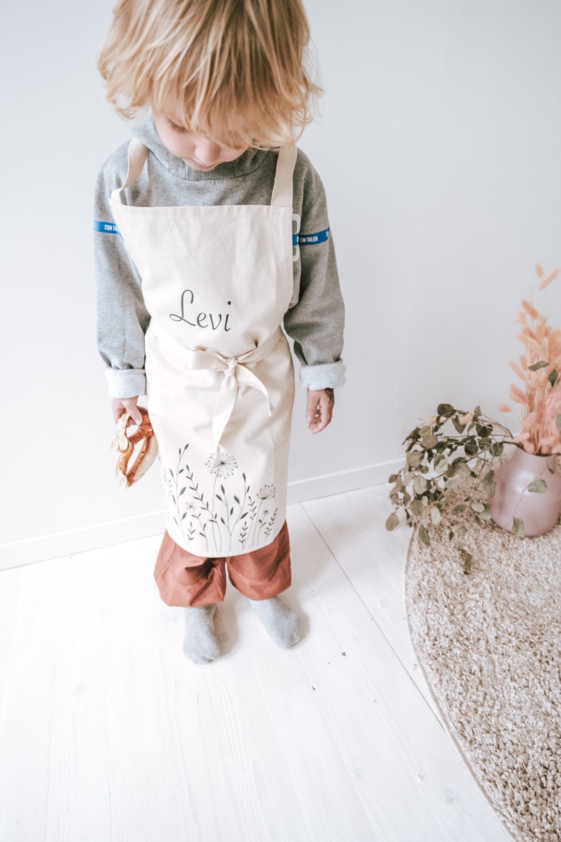 Kind mit personalisierter Schürze aus Bio Baumwolle in Naturfarbe mit Wildblumen und Pusteblumen-Motiv