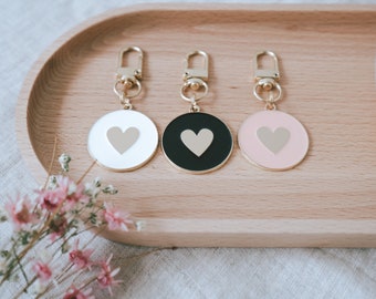 May include: Three gold keychains with round metal charms. Each charm has a different color background: white, black, and pink. Each charm has a gold heart in the center.