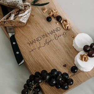 K&ouml;nnte beinhalten: Holzschneidebrett mit der Aufschrift "HAND IN HAND ein Leben lang". Das Brett ist mit Trauben, Waln&uuml;ssen und Ziegenk&auml;se dekoriert. Ein Brotlaib und ein Messer sind ebenfalls vorhanden.