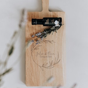May include: A wooden chopping board with a handle, engraved with the names "Julia & Finn" and the date "23 August 2025", surrounded by a floral design. A small black bottle and lavender sprigs are placed on the board.