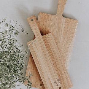 K&ouml;nnte beinhalten: Drei Holzschneidebretter mit Griffen. Die Bretter sind hellbraun mit sichtbarer Holzmaserung. Ein Brett hat den Text "DEIN TEXT" und ein Herzsymbol. Die Bretter sind unterschiedlich gro&szlig; und schr&auml;g angeordnet.