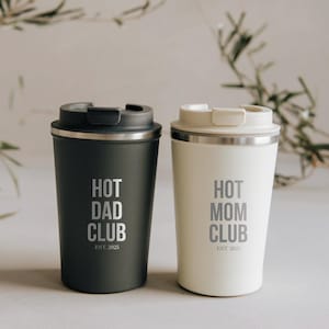 May include: Two insulated travel mugs. One is dark grey with "HOT DAD CLUB EST. 2025" printed in grey. The other is off-white with "HOT MOM CLUB EST. 2025" printed in grey. Both have black or white lids.