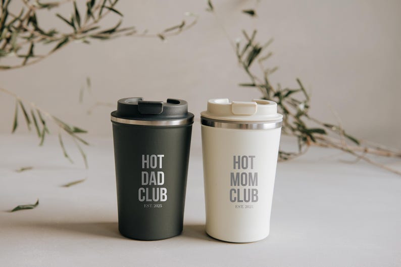 Zwei stilvolle Thermobecher mit den Aufschriften "Hot Dad Club" und "Hot Mom Club" vor einem natürlichen Hintergrund. Perfekt für Eltern, die ihren Kaffee unterwegs genießen möchten. Diese hochwertigen Becher vereinen Funktionalität und schickes Design und sind ein ideales Geschenk für stilbewusste Mütter und Väter. Holen Sie sich Ihren persönlichen Begleiter für den Alltag!