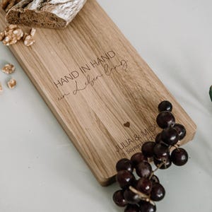 K&ouml;nnte beinhalten: Ein Holzservierbrett mit dem eingravierten Text "HAND IN HAND in Leben lang" und einem Herzsymbol. Das Brett wird von Trauben, Waln&uuml;ssen, Brot und Olivenzweigen begleitet. Das Brett tr&auml;gt auch die Namen "JULIA & MICA" und ein Datum.