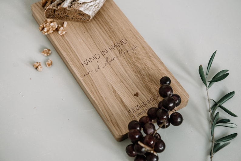 K&ouml;nnte beinhalten: Ein Holzservierbrett mit dem eingravierten Text "HAND IN HAND in Leben lang" und einem Herzsymbol. Das Brett wird von Trauben, Waln&uuml;ssen, Brot und Olivenzweigen begleitet. Das Brett tr&auml;gt auch die Namen "JULIA & MICA" und ein Datum.