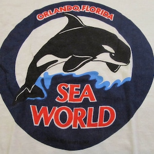 May include: A white t-shirt with a blue circle featuring a black and white orca jumping out of the water. The text "ORLANDO, FLORIDA" is above the orca and "SEA WORLD" is below the orca.