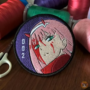 Op de afbeelding: Een ronde, geborduurde patch met een anime-personage met roze haar, groene ogen en rode markeringen op haar gezicht. Het personage draagt een rood-witte outfit. De patch heeft een paarse achtergrond en het nummer "002" in witte tekst.