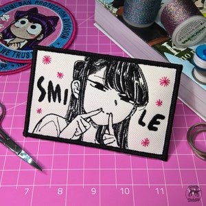Op de afbeelding: Een witte geborduurde patch met een zwart-witte illustratie van een meisje met lang zwart haar dat haar mond bedekt met haar hand. De tekst "SMILE" is in zwart geborduurd aan de linkerkant van de patch en de tekst "LE" is in zwart geborduurd aan de rechterkant van de patch. Er zijn roze geborduurde sterren rond de illustratie.