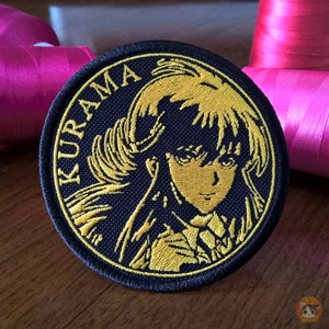 Puede incluir: Un parche bordado en negro y dorado con un personaje de pelo largo y fluido y la palabra "KURAMA" escrita en un círculo alrededor del personaje.