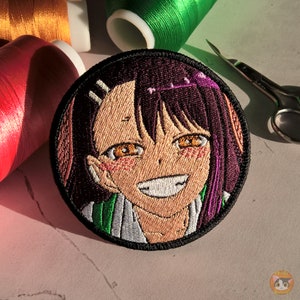 Puede incluir: Parche bordado con un personaje de anime sonriente con cabello morado y rosa, que lleva un atuendo verde y blanco. El parche es circular y tiene un borde negro.