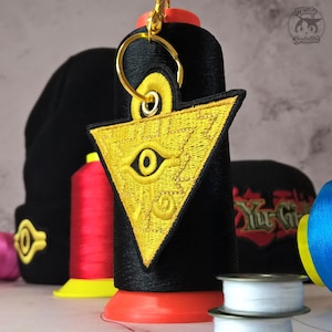 Puede incluir: Un llavero bordado en oro y negro con un diseño triangular y un ojo. El llavero está unido a un anillo dorado. El fondo incluye carretes de hilo y un gorro negro.