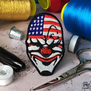 Puede incluir: Parche bordado con un diseño de cara de payaso con un motivo de bandera estadounidense. El parche tiene una combinación de colores rojo, blanco y azul, con estrellas y rayas. Rodeado de carretes de hilo, un dedal, tijeras y un rotulador.