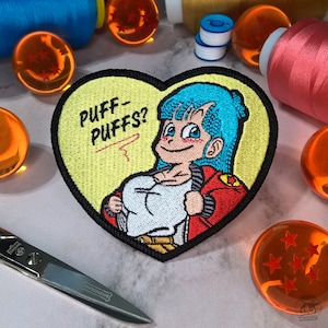 Puede incluir: Un parche bordado en forma de corazón con un personaje de dibujos animados con cabello azul, una chaqueta roja y una camisa blanca. El texto "PUFF-PUFFS?" está bordado en rojo sobre un fondo amarillo.