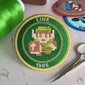 May include: A green and gold embroidered patch featuring the character Link from the Legend of Zelda video game. The patch has the text "LINK" and "1986" on it.