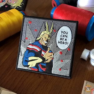 May include: An embroidered patch featuring a cartoon character with spiky blond hair and a determined expression. The character is wearing a blue, red, and white costume and is holding a fist up in the air. The patch has a gray background with red stars and a speech bubble that says "You can be a hero!"