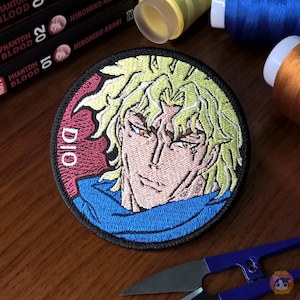 Può includere: Un cerotto rotondo ricamato con un ritratto ravvicinato di Dio Brando dalla serie anime JoJo's Bizarre Adventure. Il cerotto ha un bordo nero, uno sfondo rosa e la parola "DIO" scritta in bianco sul lato sinistro.