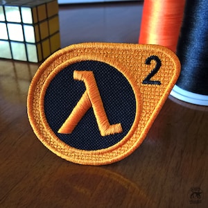 Peut inclure: Patch brodé orange et noir avec le logo Half-Life 2. Le logo est une lettre A stylisée à l'intérieur d'un cercle.