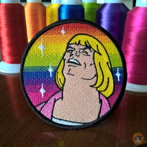 Puede incluir: Parche bordado con un personaje de dibujos animados con cabello rubio y una camisa rosa, sobre un fondo de arcoíris con estrellas blancas.