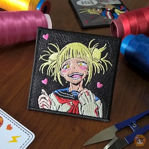 Puede incluir: Parche bordado con un personaje de dibujos animados con cabello rubio, mejillas rosadas y una sonrisa traviesa. El personaje lleva un uniforme escolar blanco y negro con un lazo rojo. El parche está rodeado por un borde negro y tiene corazones rosas alrededor del personaje.