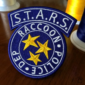 Könnte beinhalten: Ein blauer und weißer gestickter Aufnäher mit dem Text "S.T.A.R.S. Raccoon City Police Dept." und drei gelben Sternen.