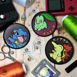 Puede incluir: Tres parches bordados para planchar con personajes de Pokemon. Los parches son circulares y presentan un Pokemon verde, azul y amarillo sobre un fondo oscuro. Los nombres japoneses de los Pokemon están escritos en los parches.
