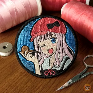 Puede incluir: Parche bordado con una chica de anime de dibujos animados con cabello rosado, usando un sombrero rojo con un lazo negro y sosteniendo un objeto marrón. El parche tiene un fondo azul con costuras blancas.