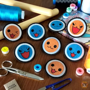 May include: Eight colorful embroidered patches with various cartoon faces. The patches are circular and have a white border with a black outline. The faces are all different colors and have different expressions. The patches are arranged in a circle on a wooden surface.