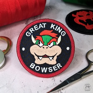 Puede incluir: Un parche bordado de color rojo, blanco y azul con el texto "Great King Bowser" y una imagen de Bowser, un personaje de la serie de videojuegos Super Mario Bros.