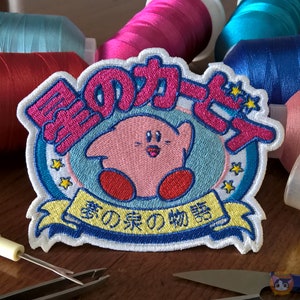 Puede incluir: Parche bordado con un personaje de dibujos animados rosa con pies rojos y una cara sonriente, sobre un fondo azul claro. El parche incluye texto japonés en rosa y una pancarta amarilla con más texto japonés. El parche está rodeado de costuras blancas.