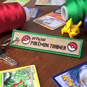 Puede incluir: Un llavero de color beige y verde con el texto "official POKEMON TRAINER" y una Pokébola roja y blanca en cada extremo.