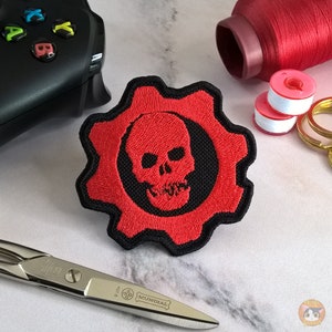 May include: Red and black embroidered patch featuring a skull inside a gear. The skull is red and the gear is black.