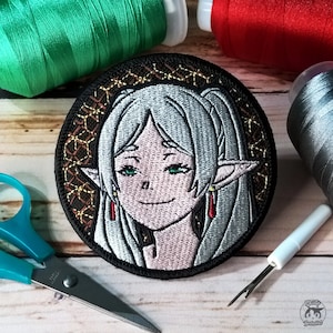 Puede incluir: Parche bordado con un personaje de pelo blanco con orejas de elfo, que lleva un collar rojo y dorado. El personaje tiene ojos verdes y una suave sonrisa. El parche es circular y tiene un fondo negro con un patrón dorado y marrón.