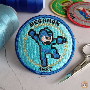 Puede incluir: Un parche bordado azul y dorado con un personaje de robot azul pixelado con el texto "MEGAMAN 1987".