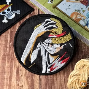 Puede incluir: Parche bordado circular que representa a un personaje de la serie de anime One Piece. El parche tiene un fondo negro con una imagen detallada del personaje con un sombrero de paja, con detalles rojos y blancos. También es visible otro parche con una calavera y huesos cruzados.