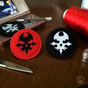 Puede incluir: Dos parches bordados con un símbolo blanco y negro sobre un fondo rojo y negro. El símbolo es una calavera estilizada con alas y una corona.