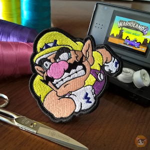 Op de afbeelding: Een geborduurde patch met een cartoonfiguur met een snor en een kroon. Het personage draagt een wit shirt met een bliksemontwerp en een paarse vest. De tekst "WARIOLAND 4" is zichtbaar op de achtergrond.